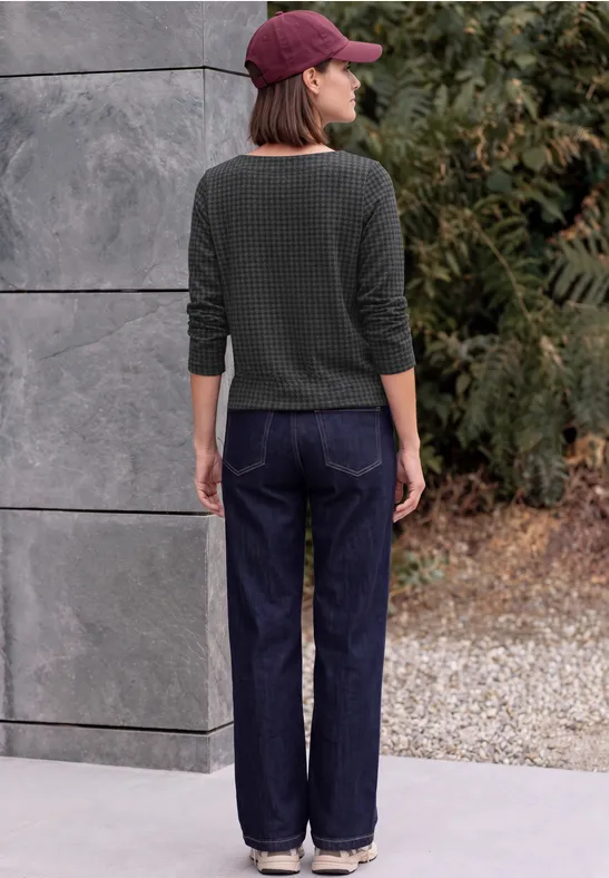 Person steht vor Wand, trägt grünen Pullover, Jeans und rote Mütze.
