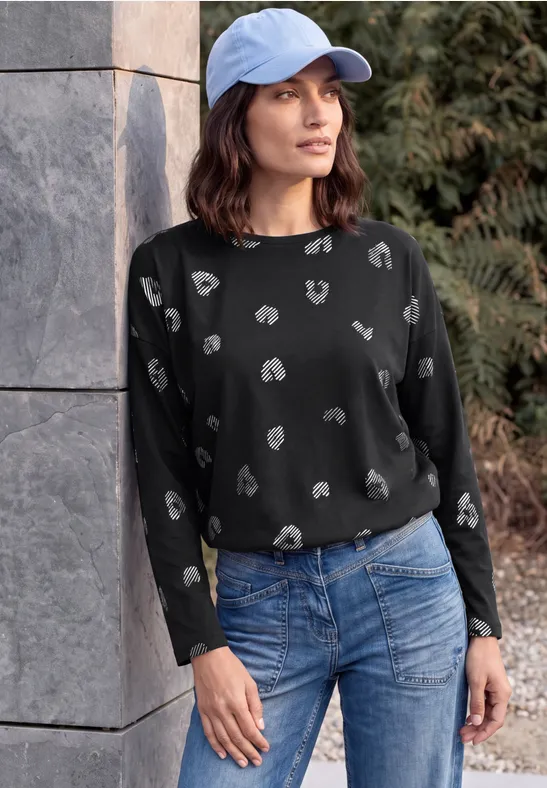 Eine Frau lehnt lässig an einer Mauer, trägt einen schwarzen Pullover mit weißem Muster, Jeans und eine blaue Kappe.