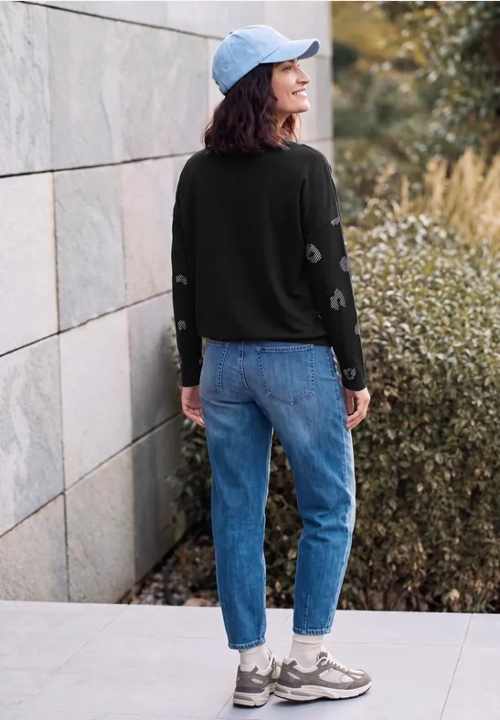 Junge Frau in Jeans, schwarzem Pullover und Cap steht vor einer Steinmauer neben Büschen.