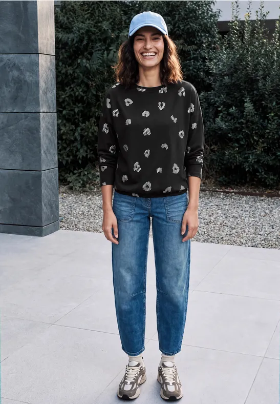 Frau steht lächelnd draußen vor modernem Bauwerk, trägt Cap, schwarz gemusterten Pullover, Jeans und Sneaker.