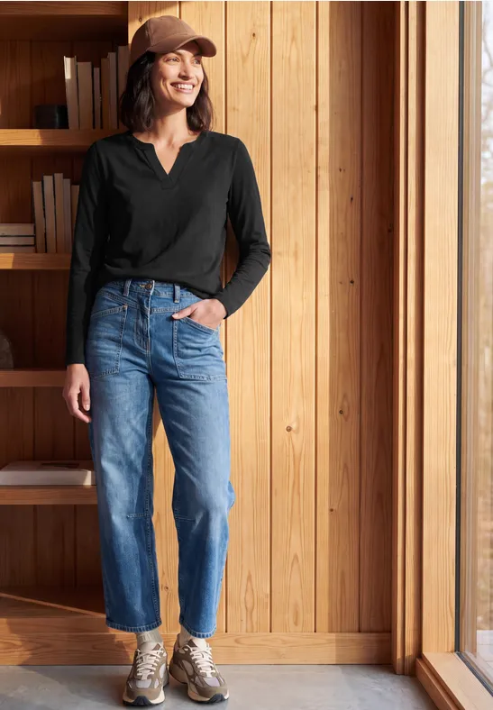 Frau mit Baseballcap lehnt am Bücherregal neben Fenster, trägt schwarzen Pullover, blaue Jeans und Turnschuhe.