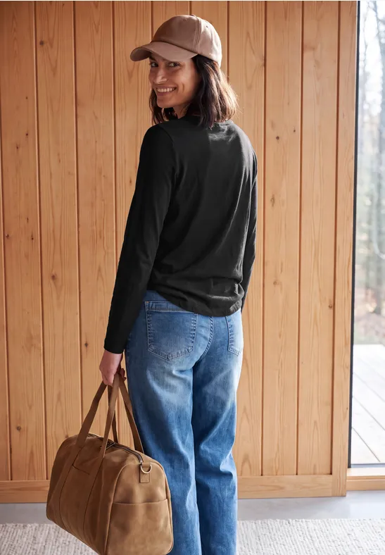 Eine Frau in Jeans, schwarzem Langarmshirt und Basecap trägt eine Tasche und lächelt vor einer Holzwand.