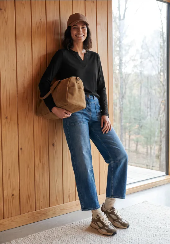 Frau in Jeans, dunkler Jacke und Cap steht mit Beuteltasche lächelnd an Holzwand neben Fenster.