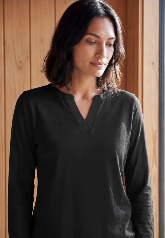 Eine Frau in schwarzem Langarmshirt steht vor Holzpaneelen und blickt nachdenklich zur Seite.