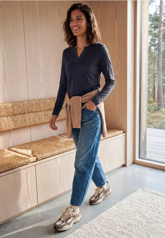 Frau in Jeans, dunkelblauem Shirt und Sneakern geht in modernem, hellem Raum an einer Holzbank vorbei.