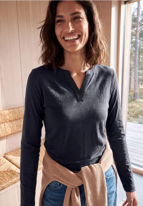 Lächelnde Frau in dunkelblauem Langarmshirt mit hellem Pullover um die Taille, in gemütlich eingerichtetem Raum.