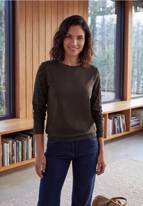 Eine lächelnde Frau in grünem Pullover und Jeans steht in einem hellen Raum vor Bücherregalen und Fenstern.