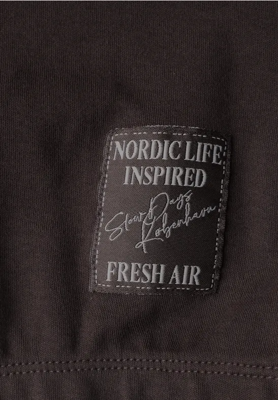 Nahaufnahme eines dunkelbraunen Kleidungsstücks mit aufgenähter Text-Plakette: ‚NORDIC LIFE INSPIRED Snow Poets FRESH AIR‘.