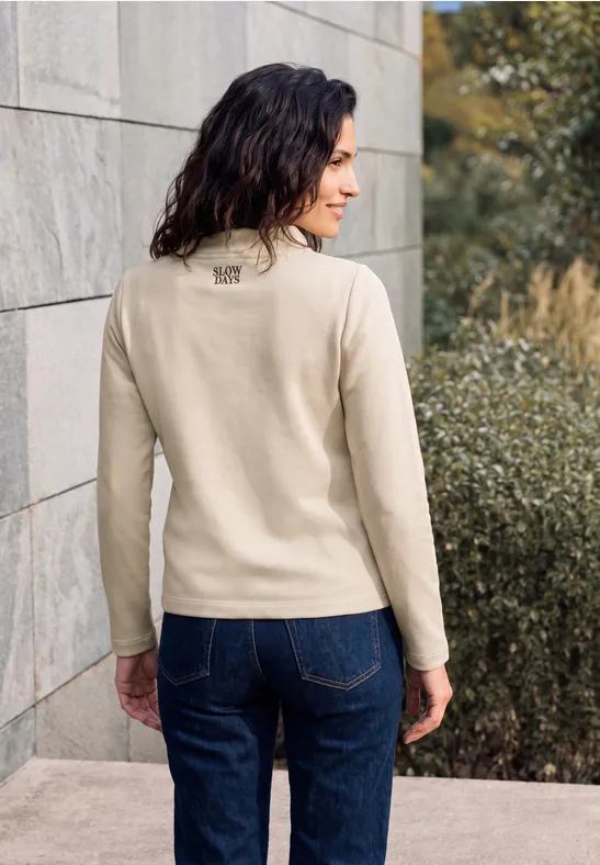 Frau steht mit dem Rücken zur Kamera, trägt hellbeige Pullover und blaue Jeans vor einer Steinmauer.