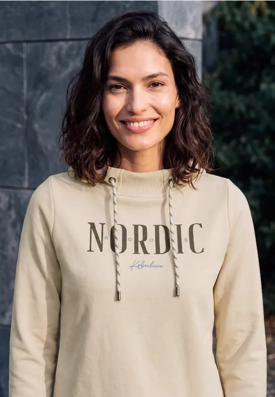 Eine junge Frau mit lockigen dunklen Haaren in einem beigen Kapuzenpullover mit 'NORDIC'-Aufdruck lächelt.