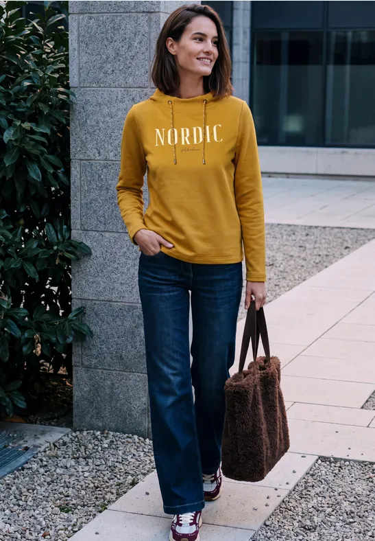 Frau im senfgelben Pullover mit Schriftzug 'NORDIC', Jeans und Tasche steht lächelnd vor moderner Fassade.