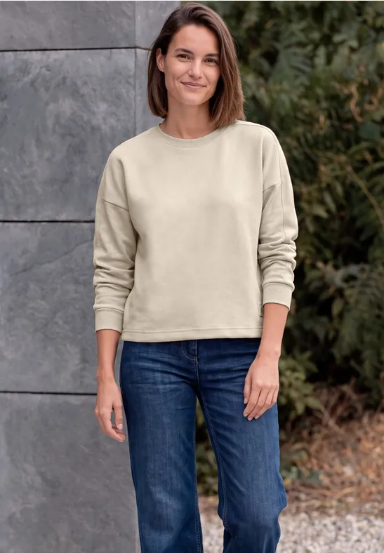 Eine lächelnde Frau in beigem Sweatshirt und Jeans steht vor einer Steinwand im Freien.