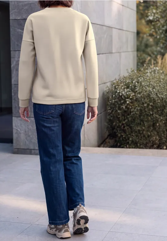 Person von hinten in beigem Pullover, blauen Jeans und Sneakers auf Pflaster neben Mauer und Sträuchern.