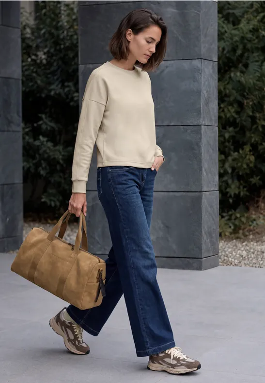 Frau mit kurzem Haar in beigem Pullover, weitem Jeans, Sneakern und großer brauner Tasche vor Steinwand.