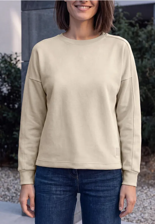 Frau in beigem Sweatshirt und blauen Jeans steht vor uns, lächelnd.