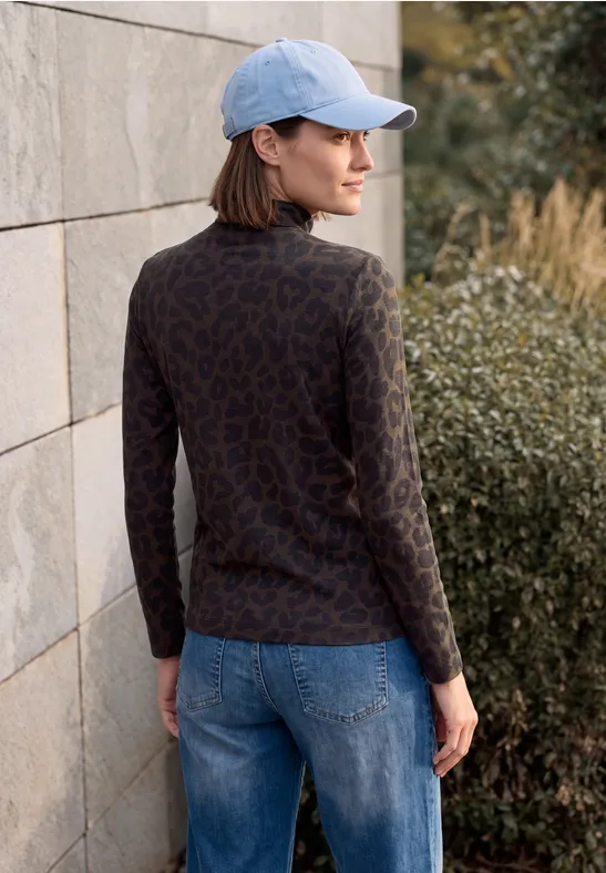 Eine Frau in Jeans, gemustertem Pullover und Cap steht an einer Mauer und schaut zur Seite.