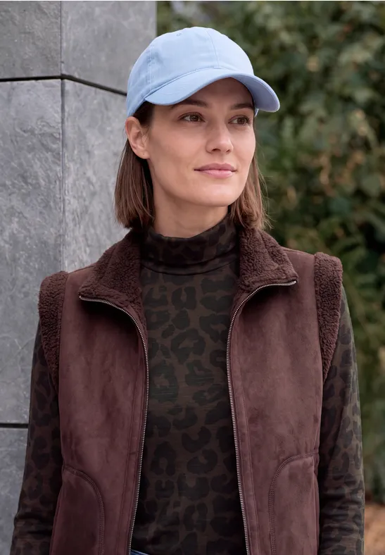Eine Frau mit blauer Baseballkappe und brauner Jacke steht draußen vor einer Mauer.