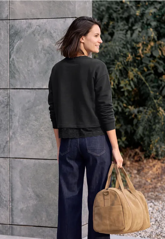 Frau von hinten in dunklem Pullover und Jeans, mit hellbrauner Tasche vor grauer Wand und Büschen.