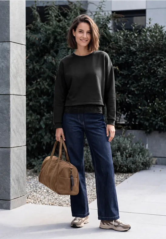 Frau in schwarzem Pullover, blauen Jeans und weißen Sneakern mit brauner Tasche steht vor Büschen.