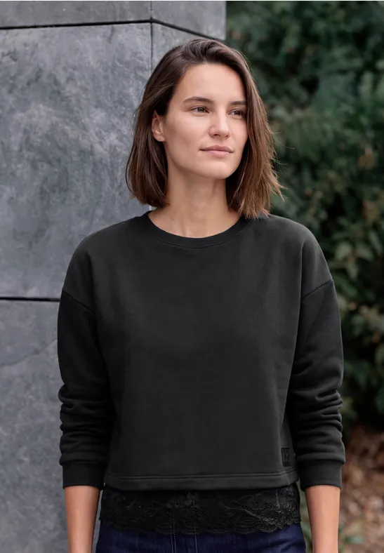 Junge Frau mit schwarzem Sweatshirt und Spitzenrock steht vor einer Steinwand und Grünpflanzen.