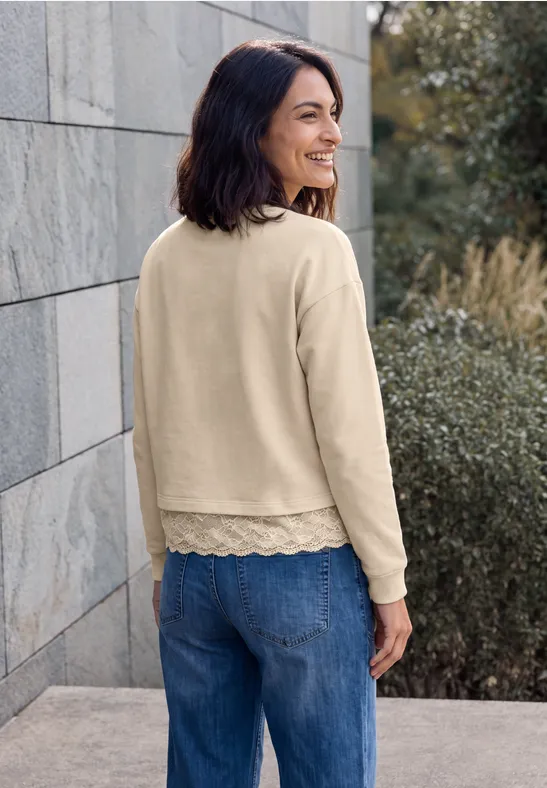 Eine lächelnde Frau in beigem Longsleeve mit Spitzenrand und Jeans lehnt an einer Steinmauer im Freien.