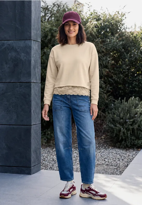 Eine Frau in Jeans, Beanie und Sneakern steht draußen vor einer Hauswand und lächelt in die Kamera.