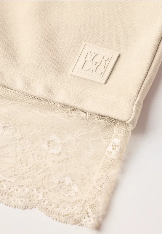 Nahaufnahme eines beigen Tops mit feiner Spitzenkante und eckigem Labeldetail.