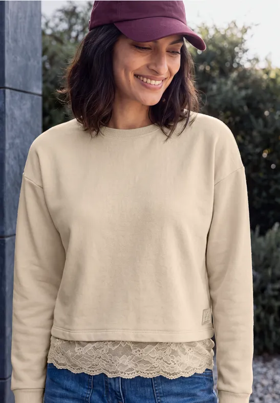 Eine lächelnde Frau in beigem cropped Sweatshirt mit Spitzenrand und violettem Cap.