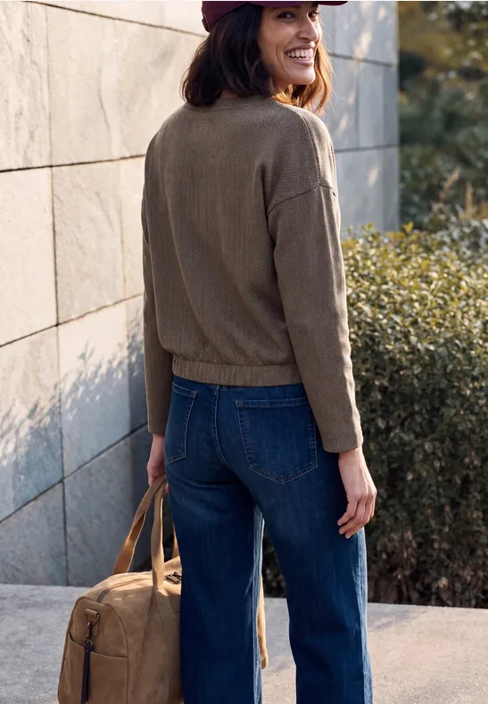 Frau im Jeans-Outfit, Jacke und Cap, trägt Tasche, steht an Wand im Freien.