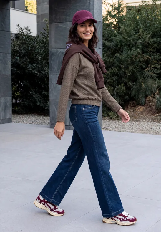 Eine Frau spaziert lächelnd im Freien in Jeans, Sneakern, Strickpullover und Kappe.