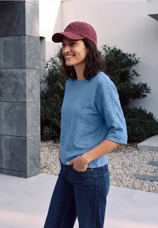 Femme souriante avec casquette rouge, t-shirt bleu et jean, debout devant un bâtiment moderne.