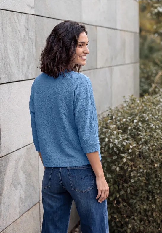 Une femme souriante en pull bleu et jean appuyée contre un mur en pierre, vue de dos, légèrement de profil.