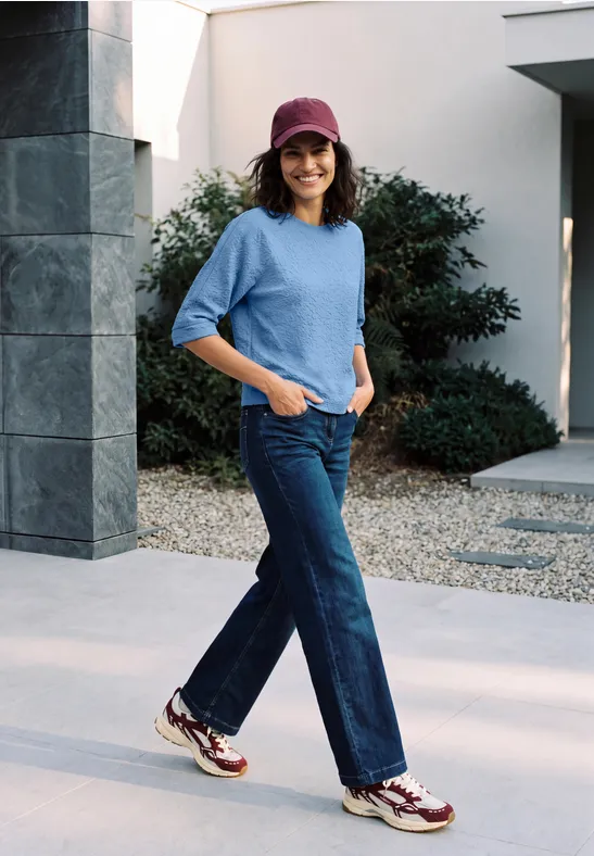 Femme décontractée avec chapeau, pull bleu et jean large se promenant devant une maison moderne.
