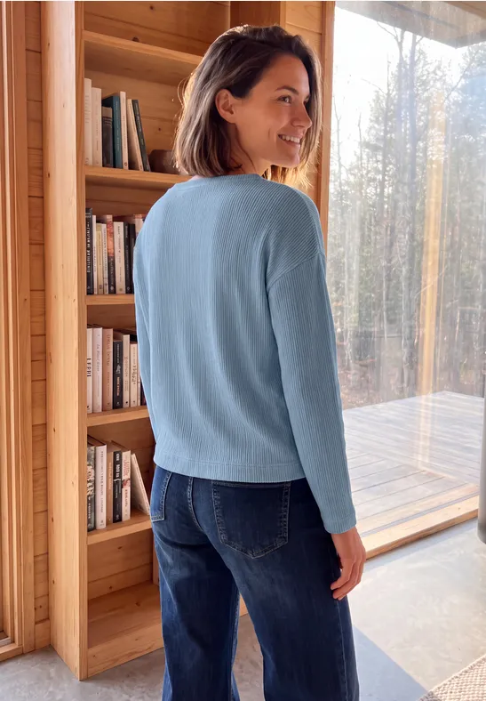 Femme en pull bleu devant une bibliothèque et de grandes fenêtres en bois.