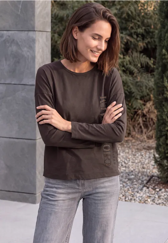 Eine lächelnde Frau mit kurzem braunen Haar, verschränkten Armen, steht vor moderner Fassade.