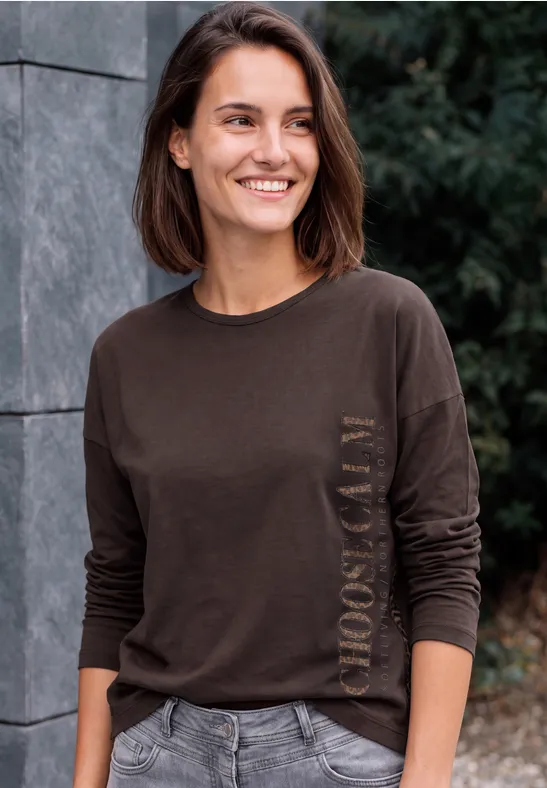 Lächelnde Frau mit kurzem braunem Haar in braunem Langarmshirt, stehend vor einer Steinwand im Freien.
