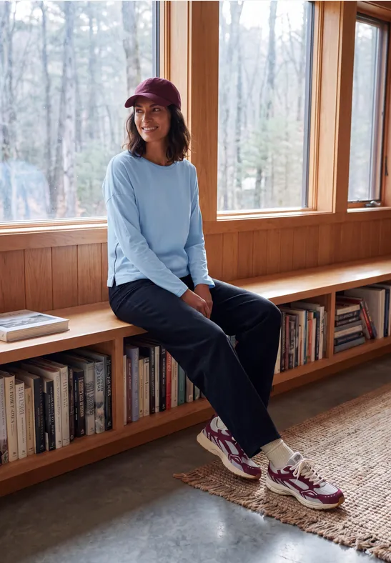 Vrouw met pet en sneakers zittend op een houten bank voor grote ramen met uitzicht op het bos.
