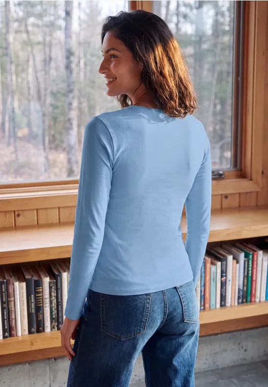 Een vrouw in een blauw hemd en spijkerbroek kijkt glimlachend bij een boekenkast en raam.