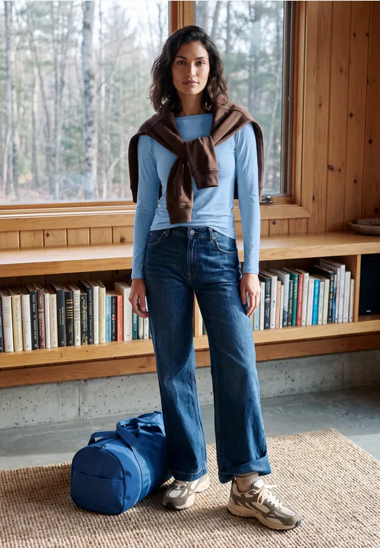 Vrouw in licht trui, jeans en sneakers met reistas, staand voor boekenplank in houten kamer.