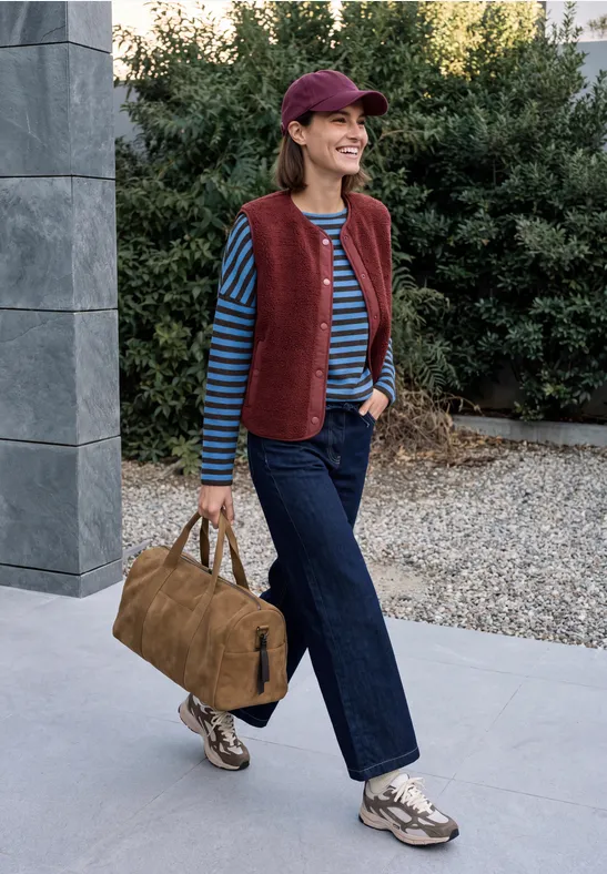 Frau in gestreiftem Shirt, roter Weste, Jeans und Cap trägt Tasche und geht draußen spazieren.