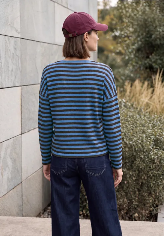 Frau von hinten in gestreiftem Pullover, Jeans und Kappe vor Steinmauer und Gebüsch.