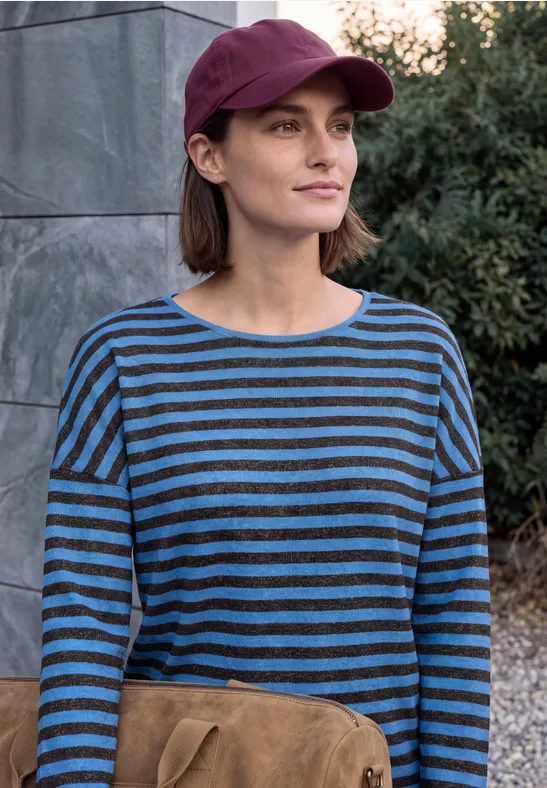 Frau im maroonen Cap posiert vor Steinmauer, trägt blau-schwarz gestreiftes Shirt.