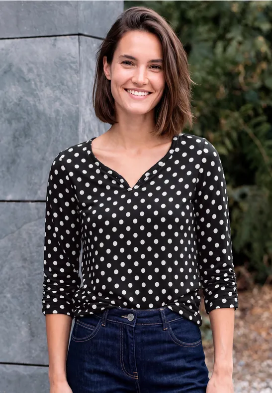 Eine lächelnde Frau in gepunkteter Bluse und Jeans steht vor einer Mauer und Pflanzen.