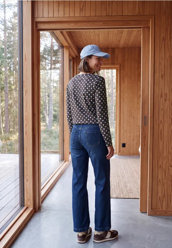 Person mit blauer Mütze, gepunktetem Pullover und Jeans steht in hellem Holzhaus, Wald draußen sichtbar.