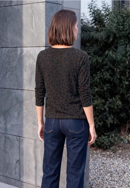 Eine Frau steht mit dem Rücken zur Kamera vor einer Steinmauer und Büschen, trägt Jeans und gemustertes Oberteil.