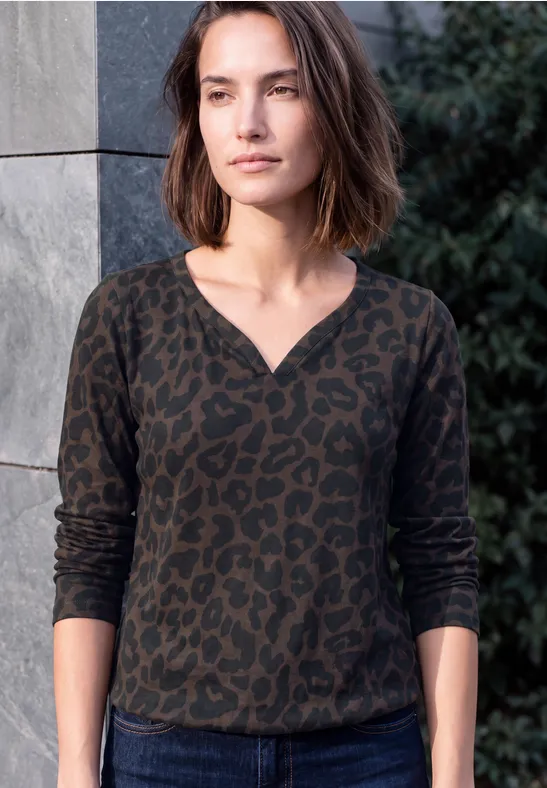 Eine Frau mit braunen Bob-Haaren trägt ein dunkelgrünes, leopardgemustertes Langarmshirt und steht vor einer Mauer.