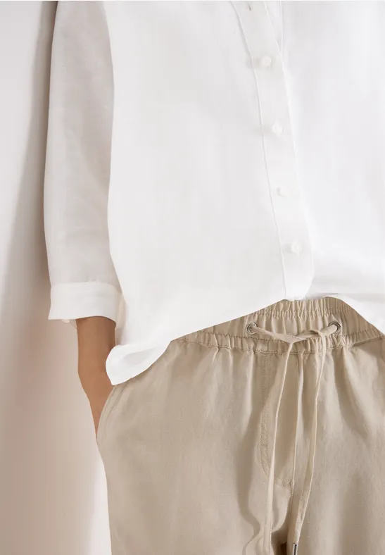 Eine Person trägt eine weiße Bluse und bequeme beige Hose, Hand in der Hosentasche.