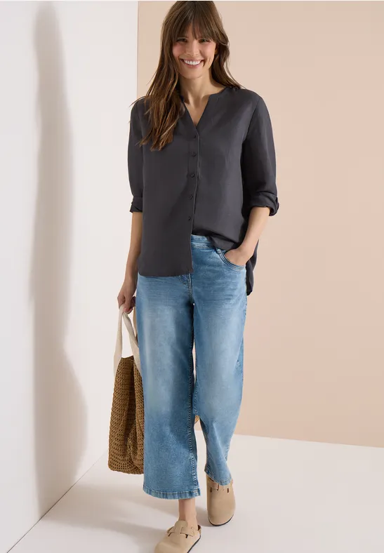 Eine Frau trägt eine dunkle Bluse mit 3/4-Ärmeln, weite blaue Jeans, beige Slipper und hält eine Korbtasche.