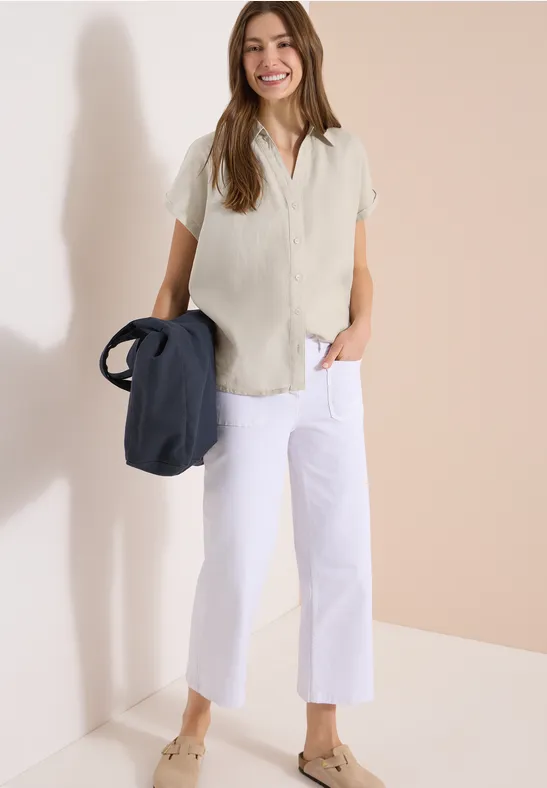 Frau trägt beiges lässiges Hemd, weiße Culottes und hält eine dunkelblaue Tasche, steht vor hellem Hintergrund.