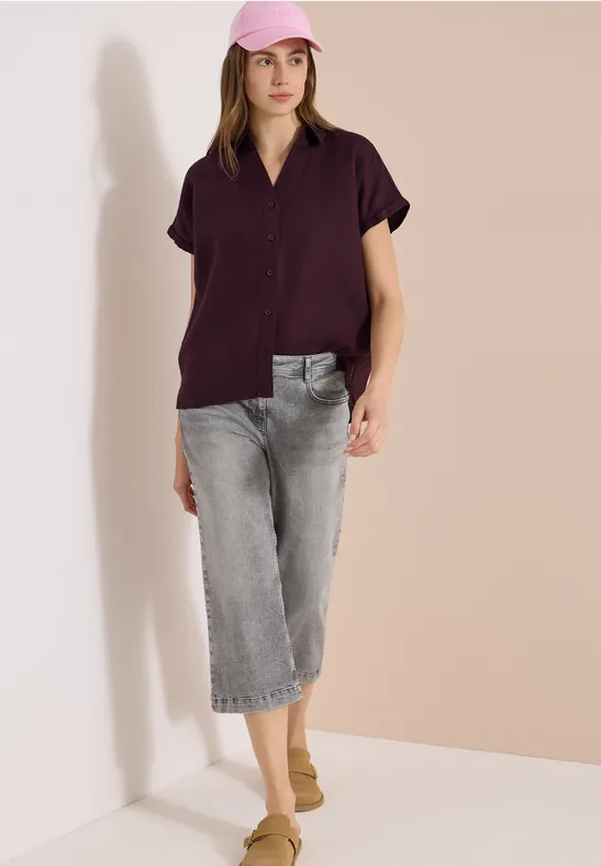 Eine Frau trägt ein kurzärmeliges, dunkelviolettes Hemd, graue Cropped-Jeans und beige Slipper vor hellem Hintergrund.